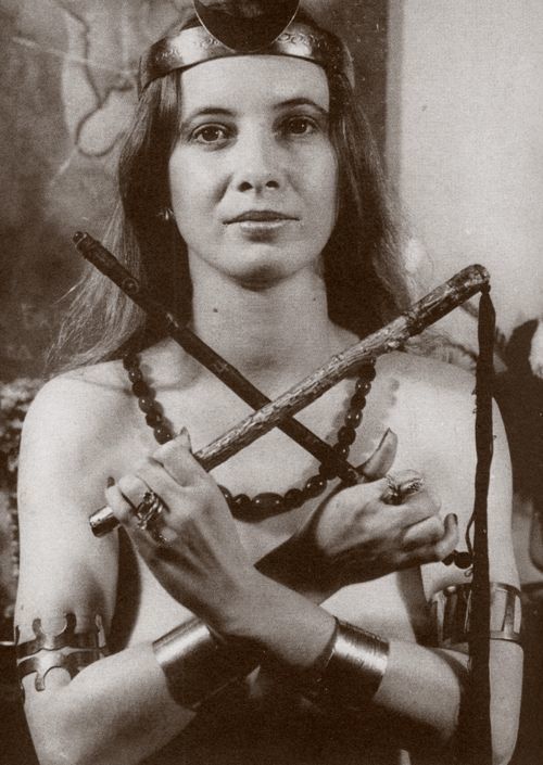 a young woman with straight hair looks at the camera, holding a flail with crossed hands