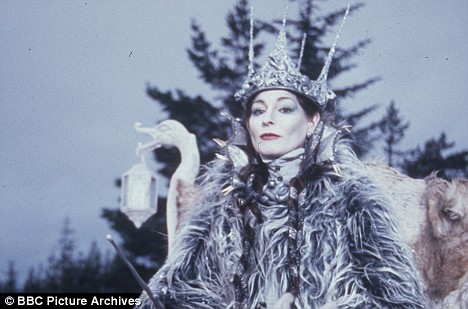 a photograph of an actress in costume as the White Witch from the classic BBC Narnia series. She has unnaturally white skin, and blue eye-shadow. She wears a crown with spikes like icicles, and a grey fur cape. Her hair is brown and in two long plaits. Behind her, there are winter trees and a grey sky, and she is sat on a carriage with a lantern. She stares imperiously at the viewer.