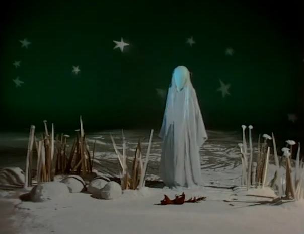 a screencap from a television program. A figure in a white robe with a covered face stands in front of a studio backdrop, with painted stars on it. There is snow on the ground and spiked grass and mushrooms around her as if dead in the winter. On the ground by her feet is the shape of a dead red squirrel.