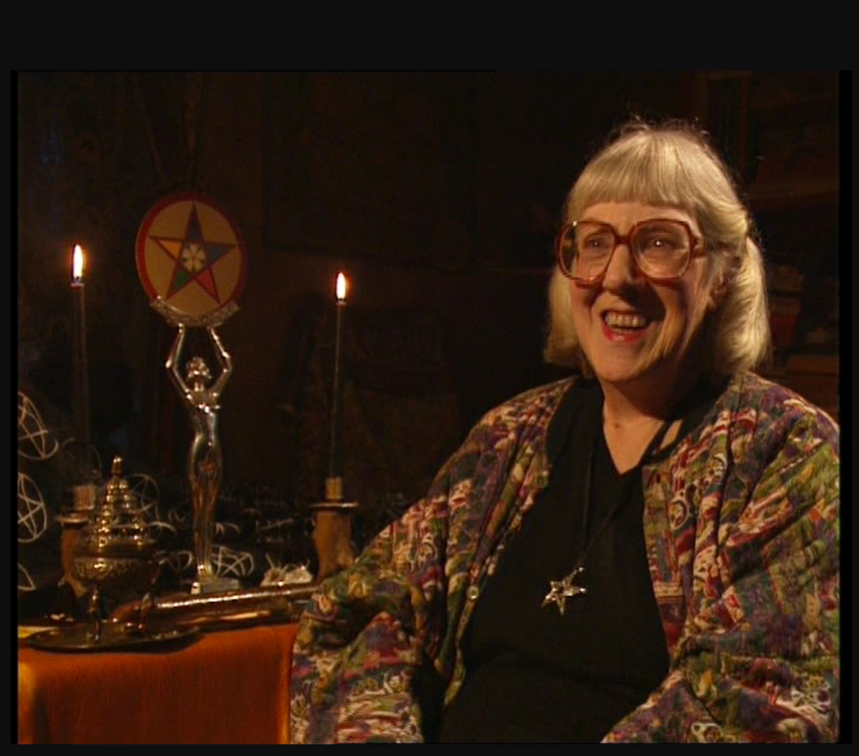 an older woman in a good jumper smiles while talking in front of a table of witchcraft tools
