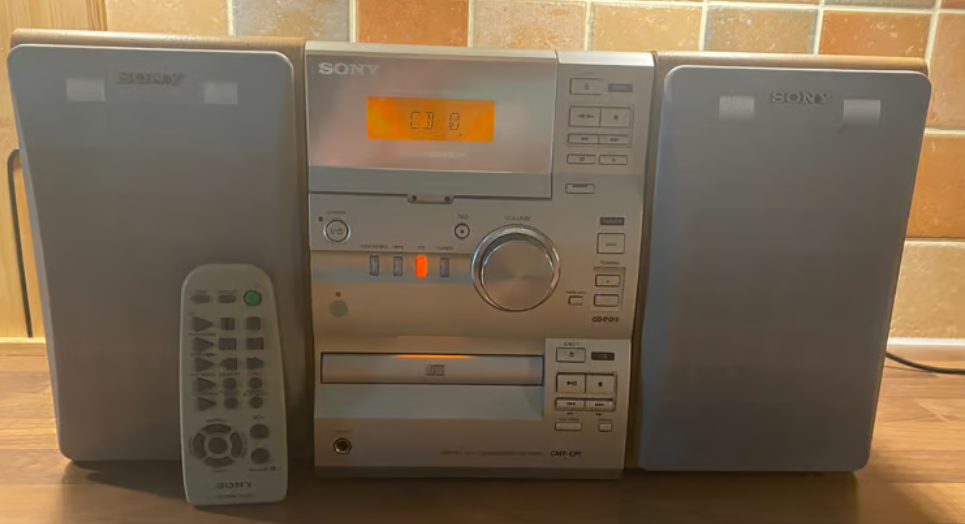 a 2000s style hi fi system, a central block with a CD player, casette player and radio,and  a box speaker either side, sat on a table